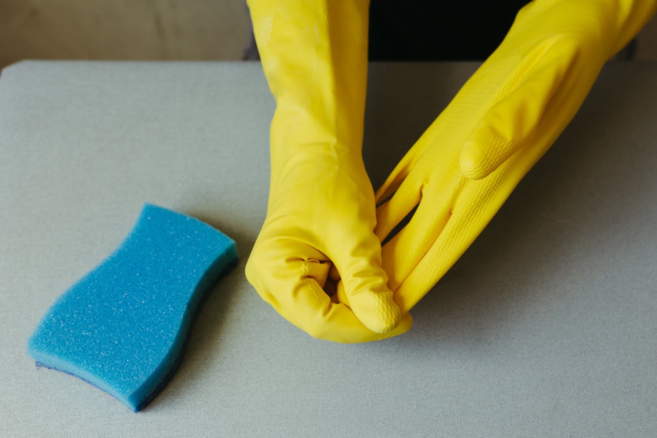 expertise-01 Close-up of hands wearing yellow gloves next to blue sponge on gray surface.